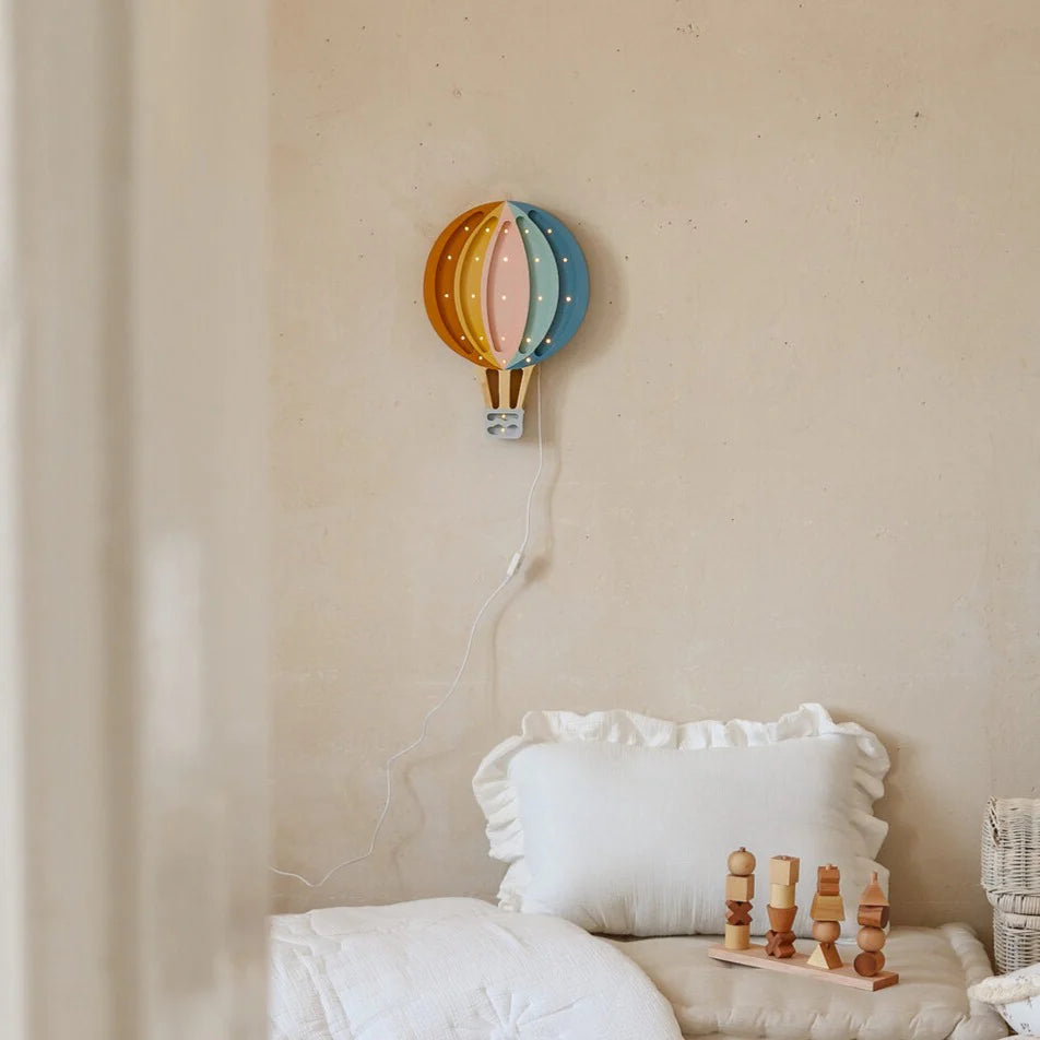 A cozy bedroom with a white pillow and blanket, three natural pinewood toys on the bed, and a Handmade Wooden Lamp - Hot Air Balloon mounted above on a beige wall.