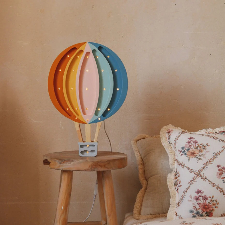 The Handmade Wooden Lamp - Hot Air Balloon sits on a pinewood stool by a cream sofa with floral, fringed pillows against a beige textured wall, adding colorful, whimsical charm to your nursery decor.