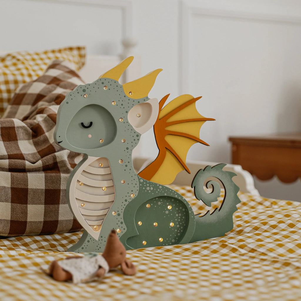 A Handmade Wooden Lamp - Dragon, Hunter Green sits on a checkered bedspread beside a plush toy and a brown-and-white checkered pillow, adding charming nursery decor to this cozy bedroom setting.