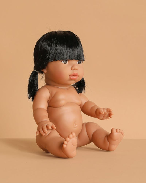 The Minikane Baby Doll (13") - Latika sits with legs outstretched and arms raised on a beige surface against a matching background. This realistic, anatomically correct doll with black pigtails is made in Spain. Clothing sold separately.