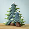 Two wooden figurines from the Snowy Fir Tree Set are elegantly painted in green with white snow accents, standing gracefully on a wooden surface. The larger tree stands behind the smaller one, creating a layered effect against the solid light blue background.