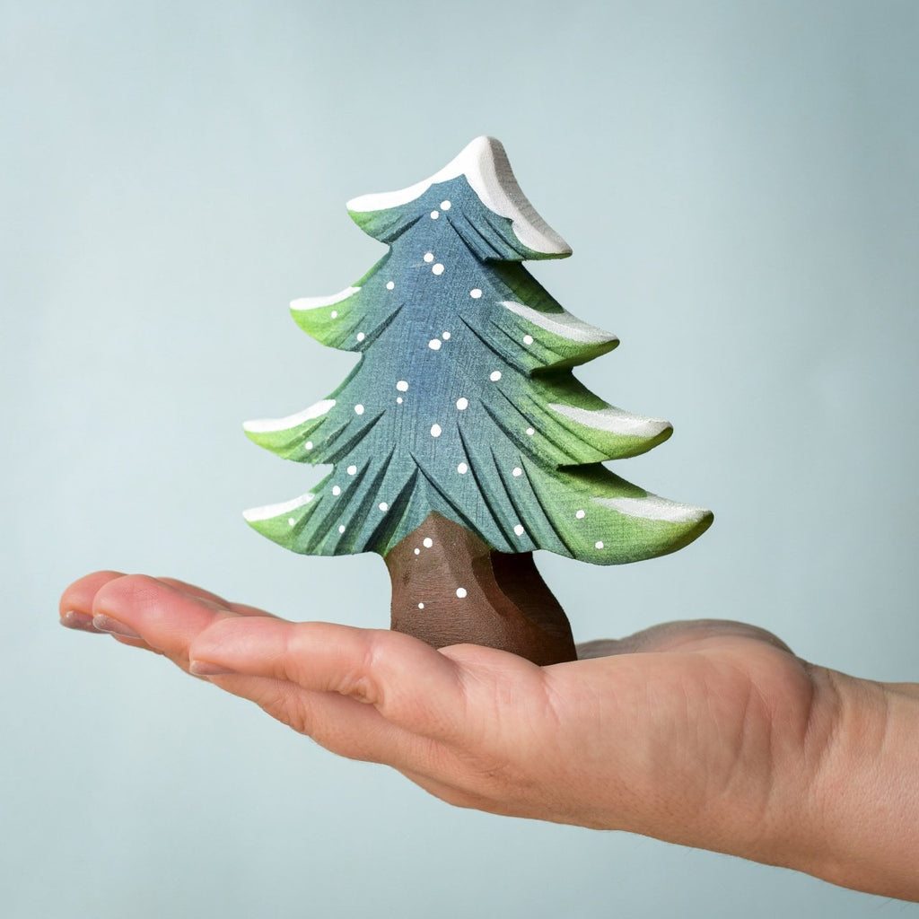 A hand holds a small, painted fir tree from the Bumbu Handmade Wooden Snowy Fir Tree Set, crafted from sustainably sourced wood and featuring snow-dusted branches, shown against a plain light blue background.