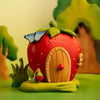 The Bumbu Handmade Wooden Strawberry House (ships in one week) is a whimsical Waldorf toy shaped like a red strawberry with a wooden door, green leaves on top, and a blue butterfly, set among grass and trees in a vibrant, playful scene.
