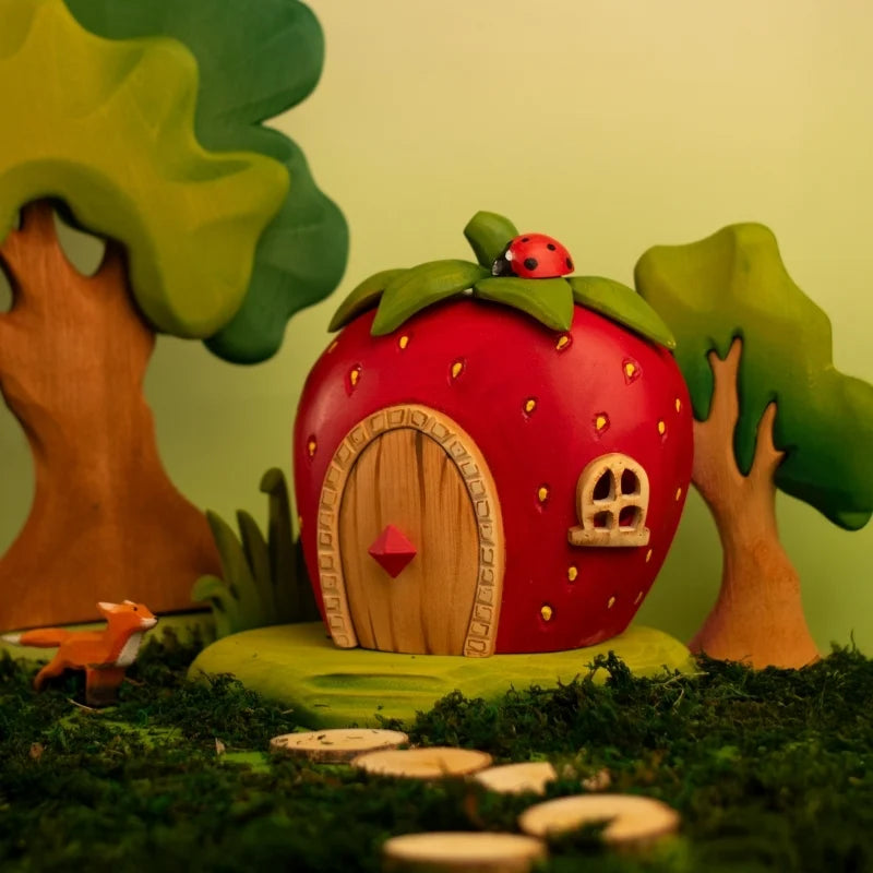 A charming scene with the Bumbu Handmade Wooden Strawberry House (ships in one week), shaped like a strawberry with door and window, surrounded by greenery. A tiny fox is on the left, while a ladybug rests atop the house.