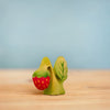 The Bumbu Handmade Wooden Strawberry Plant, inspired by Waldorf education, features a hand-painted green figure with a leaf design holding a red, yellow-dotted strawberry. Ships in one week. Displayed on light wood with a pale blue background.