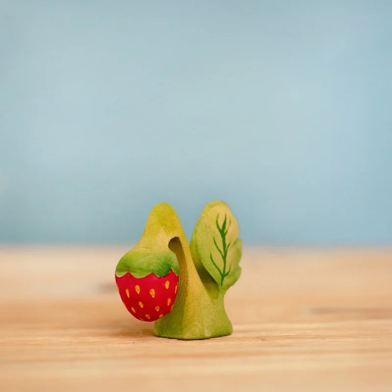 The Bumbu Handmade Wooden Strawberry Plant, inspired by Waldorf education, features a hand-painted green figure with a leaf design holding a red, yellow-dotted strawberry. Ships in one week. Displayed on light wood with a pale blue background.