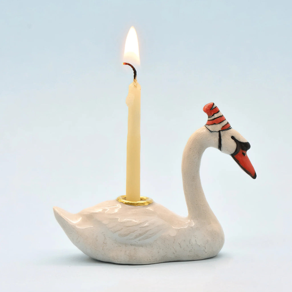 The Swan Cake Topper - Final Sale (ships in one week) is a hand-painted white ceramic swan with a red-striped party hat, holding a lit yellow candle on its back, set against a light blue background.