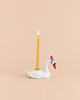 A Swan Cake Topper - Final Sale (ships in one week) features a yellow candle in a handcrafted ceramic swan holder with a red hat, glowing against a solid light peach background.