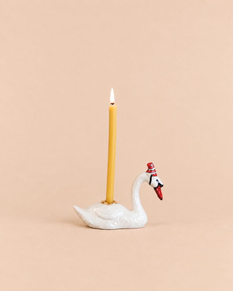 A Swan Cake Topper - Final Sale (ships in one week) features a yellow candle in a handcrafted ceramic swan holder with a red hat, glowing against a solid light peach background.