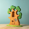 The Bumbu Handmade Wooden Ancient Oak Tree with Owl Set features green foliage, a hollow center, door, and heart-shaped window on a flat base—an inspiring playset displayed on wood with a light blue background.