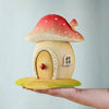 A hand displays the Bumbu Wooden Toadstool House, a handcrafted playset with a red and white spotted roof, small window, and door with a red diamond knob—crafted from sustainably sourced wood against a plain background.