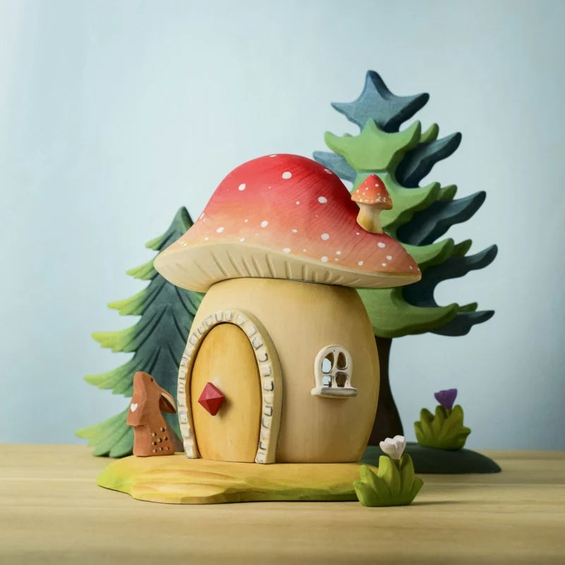 The Bumbu Wooden Toadstool House is a red-capped mushroom playset with white spots, small windows, and a round door, featuring handcrafted trees, mushrooms, and flowers made from sustainably sourced wood.