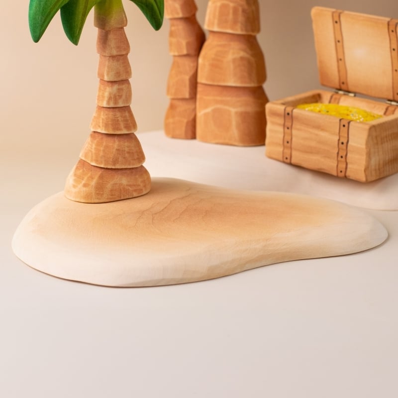 The Bumbu Handmade Wooden Treasure Hunt Set includes a palm tree, sand, rock formations, and an open treasure chest with yellow coins—all crafted in a smooth, natural style ideal for Montessori learning.
