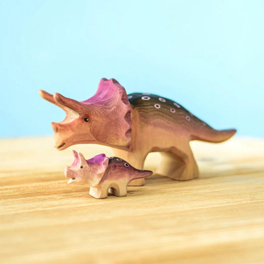 Two wooden toy dinosaurs, resembling triceratops, are displayed on a light wooden surface with a blue background. Handmade from sustainably sourced wood, one is larger with pink accents on the head and back, and the other is smaller with similar pink and purple detailing. Both are painted with non-toxic paint. This is the Bumbu Handmade Triceratops Dinosaur Set.