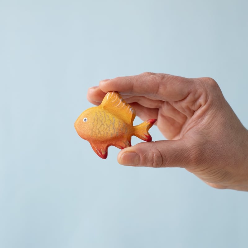 A hand holds a small, orange and yellow toy fish with red-tipped fins from the Bumbu Handmade Wooden Tropical Fish Set against a plain, light blue background.