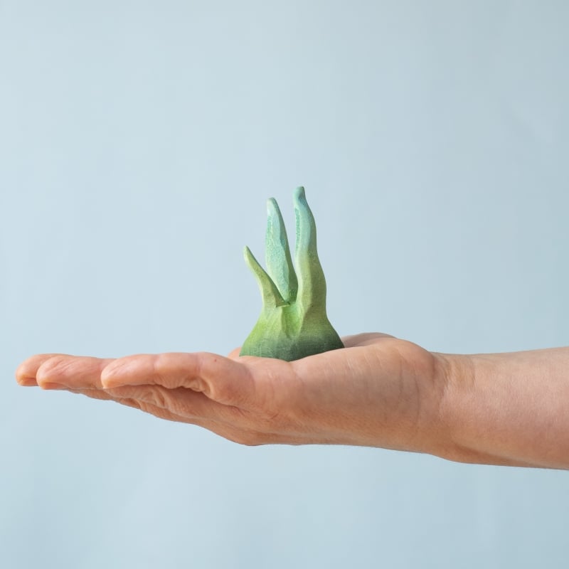 An open hand holds a small, abstract green piece from the Bumbu Handmade Wooden Tropical Fish Set—reminiscent of Montessori toys—against a pale blue background.