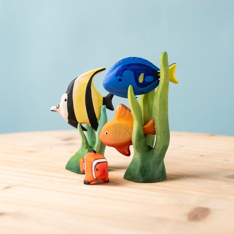 Four vibrant wooden fish from the Bumbu Handmade Wooden Tropical Fish Set stand among green seaweed on a light surface with a soft blue background—a charming addition to any Montessori toy collection.