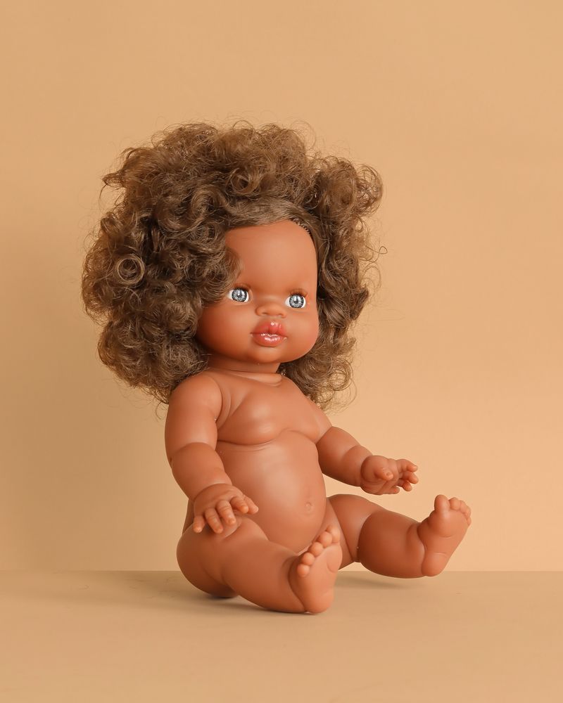 The Minikane Baby Doll (13") - Charlie, made in Spain, with brown skin, curly brown hair, and blue eyes sits on a beige surface against a beige background. Clothing sold separately.