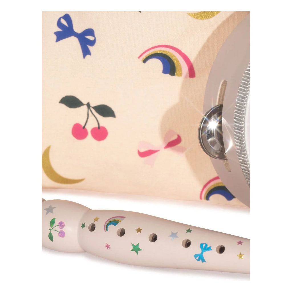 A Konges Sloejd Music Set Petite - Rainbow Twinkle wooden musical flute decorated with rainbow, star, and cherry decals, positioned in front of a patterned fabric bag with a tambourine edge.