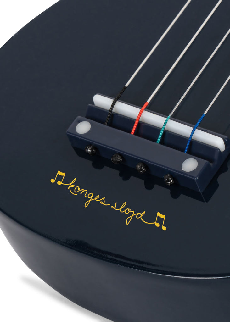 Close-up of the Konges Sloejd Wooden Ukulele - Midnight, a dark blue FSC-certified 4-string ukulele. The bridge and strings are visible, with “konges slojd” in yellow script below the bridge, flanked by musical notes.