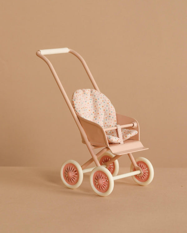 The Maileg Stroller, Micro - Powder is a small, pastel pink stroller with white wheels and a polka-dot fabric seat, shown against a beige background.