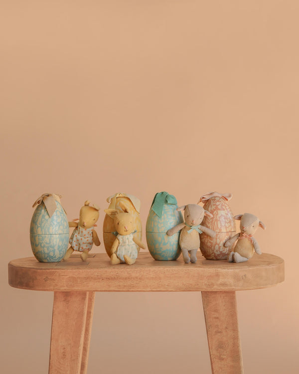 The Maileg Easter Egg & Friends Set features six pastel fabric eggs, each with a Micro Rabbit soft toy bunny, displayed on a light wooden bench against a simple beige backdrop.