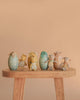 The Maileg Easter Egg & Friends Set features six pastel fabric eggs, each with a Micro Rabbit soft toy bunny, displayed on a light wooden bench against a simple beige backdrop.