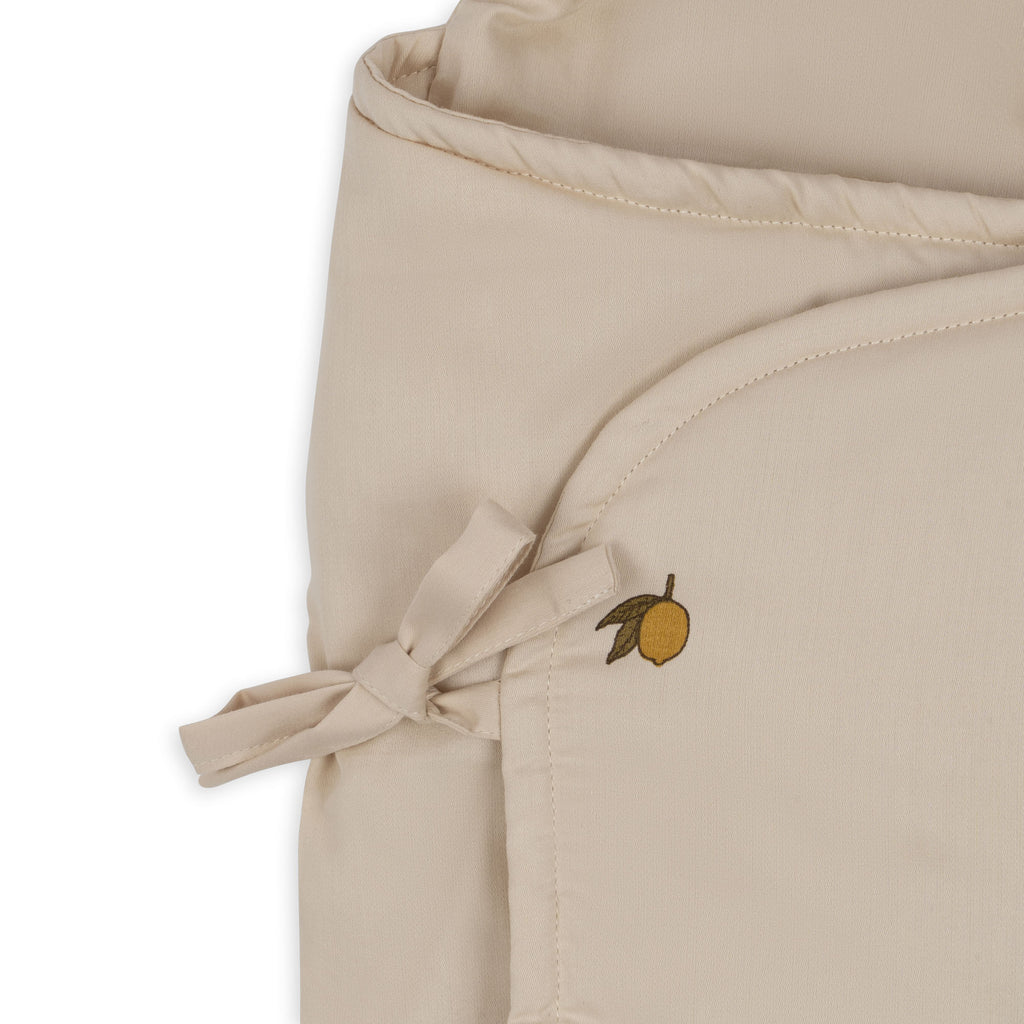 The Konges Sloejd Doll Sleeping Bag - Lemon features beige organic cotton fabric with a stitched bow and a small embroidered lemon with a leaf, offering soft padding perfect for dolls.