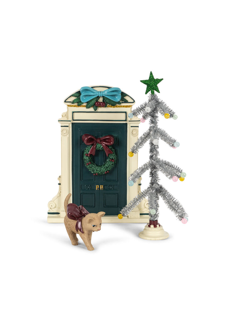A Konges Sloejd Christmas Door miniature decorated with a wreath and a blue bow, standing next to a small silver tinsel Christmas tree and a toy cat wearing a maroon bow.