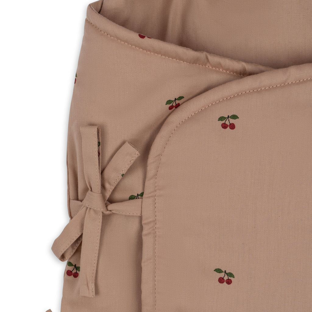 A close-up of the Konges Sloejd Doll Sleeping Bag - Cherry Blush, made from soft, light brown organic cotton with red cherry patterns, fabric ties, rounded edges, and recycled polyester filling.