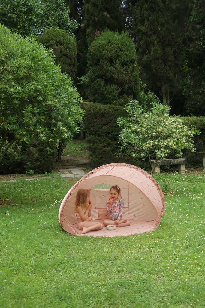 Two young children sit inside a Konges Sloejd Lace Pop-Up Beach Tent UPF 50+ on a grassy lawn, surrounded by lush green trees and bushes, sharing a drink and snacks together.
