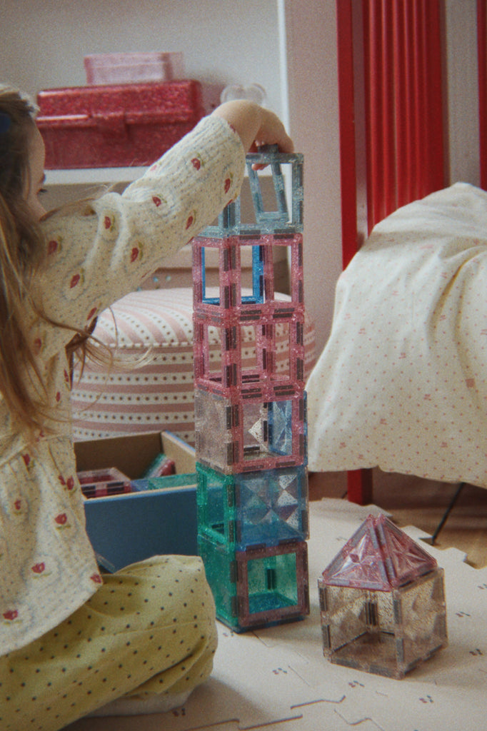 A child develops fine motor skills using the Konges Sloejd Piece By Piece Magentic Building Set to stack glittery, translucent magnetic tiles into a tall tower next to a small pyramid-shaped structure.