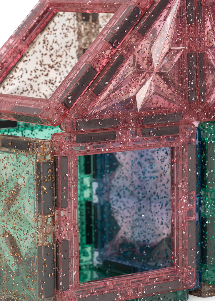 A house-shaped structure built from translucent, glitter-filled magnetic tiles in pink, green, and blue, created using the Konges Sloejd Piece By Piece Magentic Building Set.