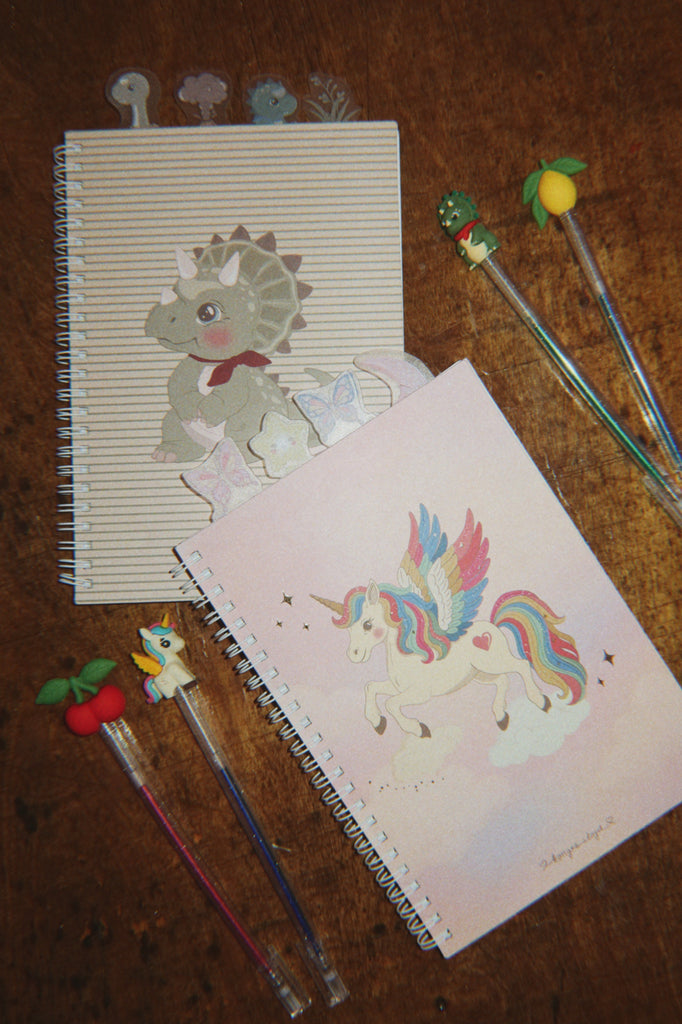 Konges Sloejd My Stationery Box - Unicorn containing two A5 notebooks featuring a dinosaur and a winged unicorn, with four glitter gel pens topped with fruit and character charms.