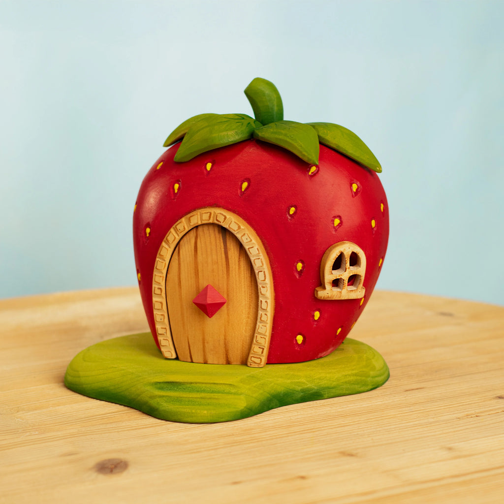 The Bumbu Handmade Wooden Strawberry House, shaped like a strawberry with a wooden door and tiny window, sits on a green base atop light wood—an enchanting Waldorf toy. Ships in one week. Pale blue background shown.