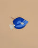 A Surgeon Fish Pencil Case, crafted from blue cotton with a white tail and fins, rests on a beige surface. A pencil peeks out from its zippered opening.