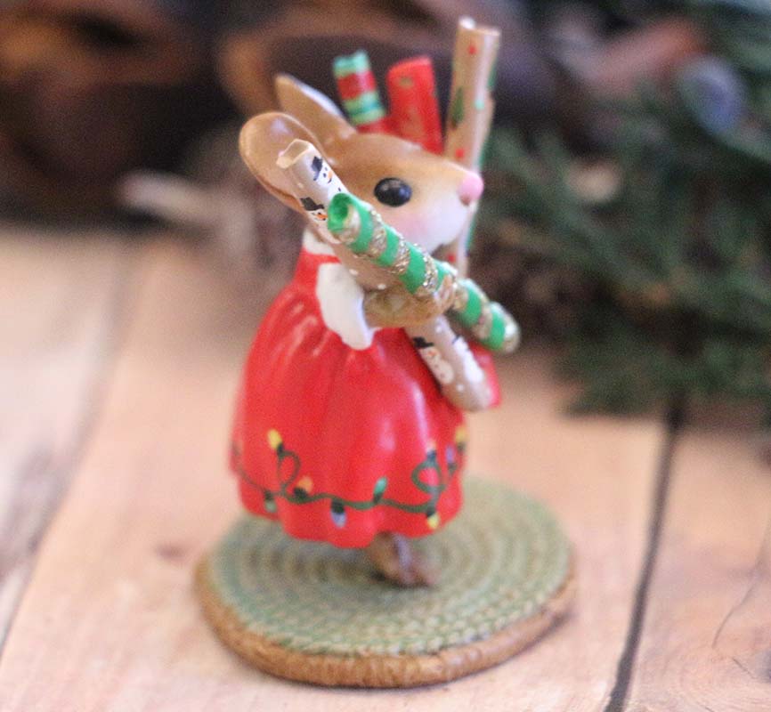 A hand-sculpted Wee Forest Folk Tiny Collectible - Wrap it Up! (Not a Toy) mouse in a red dress stands on a textured base, holding festive crackers, with blurred pine branches adding holiday charm.