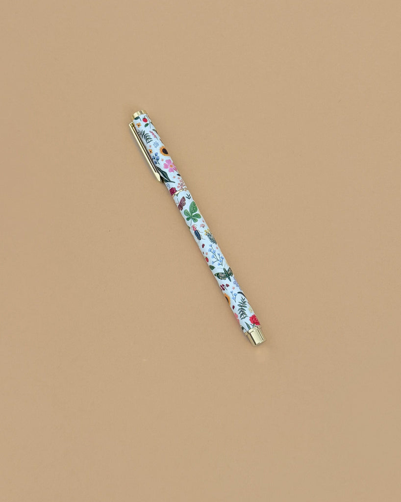 The Curio Pen, a 0.5mm gel ink rollerball with a floral design in red, green, and blue accents, rests diagonally on a plain light brown surface.