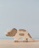 A Holztiger Triceratops stands on a light wooden surface against a pale blue background. The painted toy, made in Europe, is primarily beige with gray spots, featuring three horns and the characteristic frill of a triceratops.