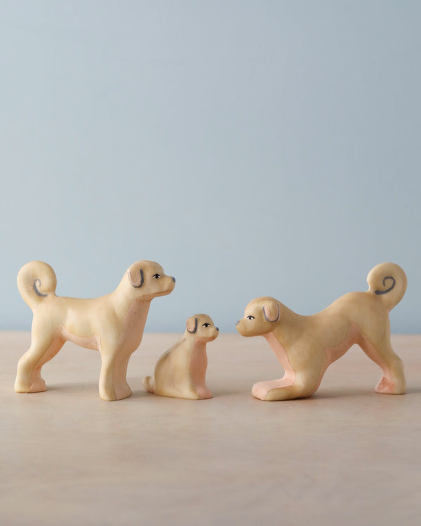 The Handmade Wooden Dog Family features three small ash wood dog figures—two adults and a puppy—displayed on a light surface with a pale blue background. One adult is in a playful pose. Eco-friendly, handcrafted toy made in Romania.