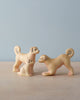 The Handmade Wooden Dog Family features three light tan ash wood dog figures with curly tails—two standing and one sitting—arranged on a light wooden surface against a pale blue background.