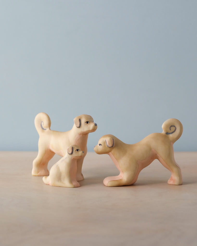 The Handmade Wooden Dog Family features three light tan ash wood dog figures with curly tails—two standing and one sitting—arranged on a light wooden surface against a pale blue background.