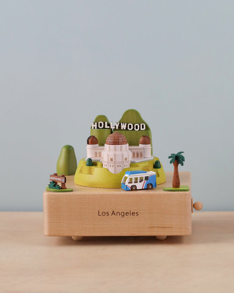 The Los Angeles Wooden Music Box showcases miniature models of iconic Los Angeles landmarks, such as the Hollywood sign, Griffith Observatory, and a blue-and-yellow bus. The base, inscribed with "Los Angeles," rests on a wooden surface, making it an ideal keepsake gift that celebrates the city's unique cultural landmarks.