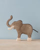 A high-quality Holztiger Elephant, Trunk Raised stands on a light-colored surface against a light blue background. Handcrafted from wood, typical of HOLZTIGER figures, the elephant is painted gray with light-colored tusks, feet, and a black tail. The figure has minimal details and a smooth finish.