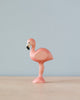 A Handmade Holzwald Flamingo figurine, painted pink, stands on one leg with a plain, light blue background.
