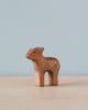A Handmade Holzwald Standing Fawn, skillfully carved with visible wood grain and decorated with small white dots on its side, stands against a soft blue background.