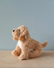 A plush toy dog with soft, light brown fur sits on a wooden surface against a sky-blue background. The toy features a white belly, black nose, and wears the iconic Steiff "Button in Ear." The product is the Steiff, Jimmy Berno Goldendoodle Puppy, 10".