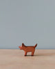 A small, high-quality Handmade Holzwald Brown Dog figurine stands upright against a plain blue-gray background. The figurine is simplistic, with minimal detailing on its body.
