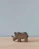 A small Handmade Holzwald Baby Rhino figurine, crafted from high-quality sustainable materials, stands on a smooth surface against a plain light blue background, showcasing its simplistic and artistic carved details.