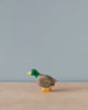 A small, high-quality Handmade Holzwald Male Duck figurine with a green head, grey body, and orange feet, standing against a plain, light blue background.